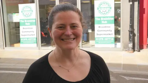 John Fairhall/BBC Laura Fowle, wearing a black round-neck top. She has dark pulled-back hair and is wearing a thin gold necklace. She is smiling at the camera, and stands in front of two signs which advertise the Braintree Hub