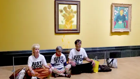 JSO Handout Three activists in white tshirts bearing the words JUST STOP OIL sit underneath Van Gogh's Sunflowers in a brown frame, which has orange soup on it that they have thrown. Another painting is on the yellow wall next to it.
