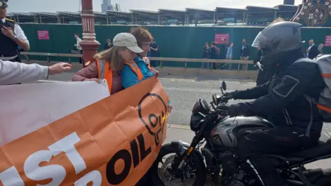 JSO Handout Protesters holding an orange Just Stop Oil banner block a motorcyclist dressed in black on a road