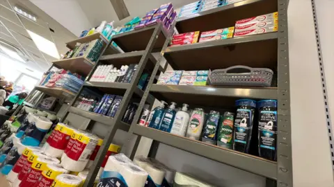 Stuart Woodward/BBC Several shelves inside the Braintree Community Supermarket. The shelves contain several different items including shampoo and handwash bottles, kitchen towels, deodorants and toothpaste. The shelves are dark grey metal in colour.