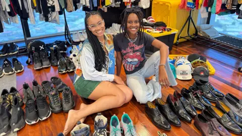 Courtesy of Aliyah Clarke Aliyah Clarke and Kaleen Sanois sell entertainter Machel Montano's clothes at pop-up shop. They sit on the floor, surrounded by pairs of shoes.