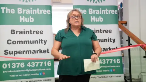 John Fairhall/BBC Tina Townsend, standing in front of two green and white signs which read "The Braintree Hub, Braintree Community Supermarket" along with a telephone number and an email address. There is a red ribbon behind Tina, and she is wearing a green shirt and black trousers