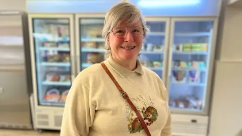 Stuart Woodward/BBC Carolyn Hobbs, stood in front of four transparent fridge doors. She is wearing a grey jumper with two hedgehogs embroidered on the front. She has a leather brown handbag strap across her chest. She has large thin-rimmed spectacles, and grey hair. She is smiling at the camera