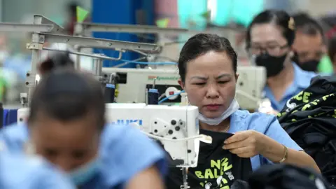 Getty Images Vietnamese garment factory workers stitch apparel at a factory in Ho Chi Minh City on April 3, 2025. They are seen in a row in light blue shirts bent over white sewing machines.