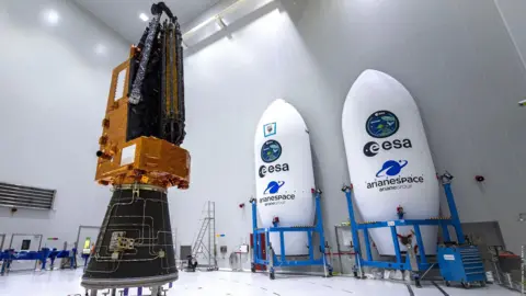 ESA-CNES-ARIANESPACE The biomass satellite sits inside an all white warehouse. It has a black cone-like base, an orange l-shaped top that has the look of foil, with the antenna attached and furled. To the left of the satellite sits two shells which will hold the satellite, they are white elongated cones with "ESA" written on them, and held in blue scaffolding trolleys.