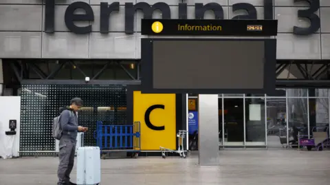 EPA-EFE/REX/Shutterstock A man stands outside Heathrow Terminal 3, looking at his phone with a suitcase beside him. An empty digital information screen is overhead, and the area is quiet with a few trolleys and barriers visible in the background.