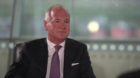 BBC Thomas Woldbye, the boss of Heathrow Airport, sitting and talking, wearing a light coloured shirt, a dark coloured jacket and a purple tie.