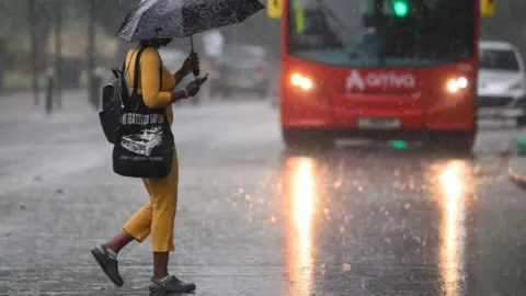 Getty Images A woman wearing a yellow top and trousers and carrying an umbrella crosses the road in the pouring rain in front of a red double decker bus