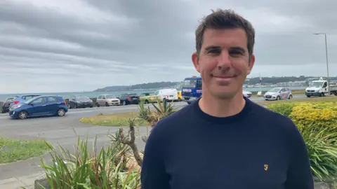 BBC Tom Pinsard is smiling at the camera as he stands on a garden. Behind him is a road which has cars parked along it next to a harbour wall with the sea extending beyond. He has short brown hair and is wearing a navy crew cut jumper.