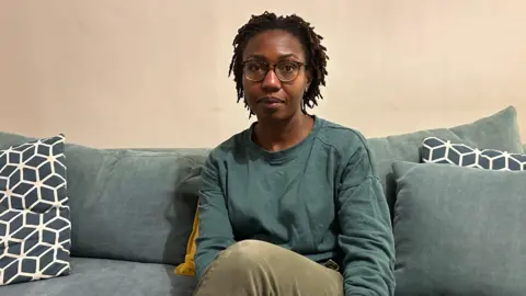 Femilola Miller A woman wearing glasses and a green jumper sits on a sofa and stares into the camera, with cushions around her and a plain wall behind.