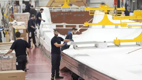 PA Media A spacious industrial workshop where several workers in dark clothing are assembling or inspecting large white objects. Yellow overhead cranes and lifting equipment are visible above. The workspace is clean and organized, with various tools and machinery around.