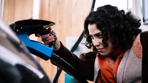 Getty Images Woman with dark hair plugs in electric vehicle to charging point