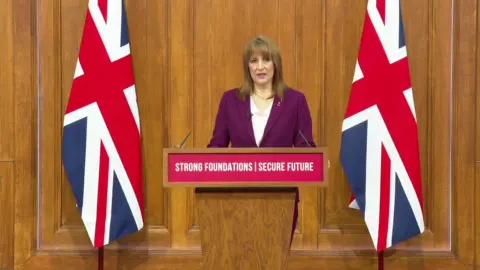BBC Chancellor Rachel Reeves giving a speech in the wood panelled Downing Street press conference suite between two union jack flags at a lecturn with the words Strong Foundations Secure Future. She is wearing a purple blazer and pale pink v neck top