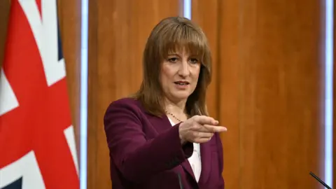 Reuters Rachel Reeves points as he takes questions from reporters, with a union jack flag in the background