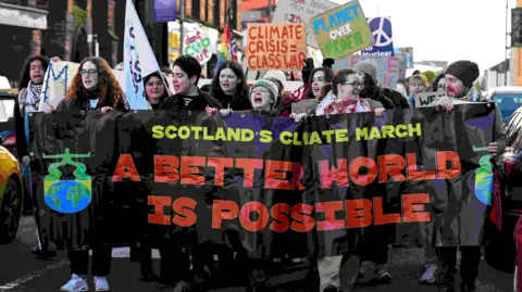 Reuters A large group of protesters marches down a street holding a wide black banner that reads “Scotland’s Climate March” and “A Better World Is Possible” in bold coloured letters. People at the front appear to be chanting, while others behind them hold signs with messages such as “Climate Crisis = Class War,” “Planet Over Profit,” and a peace symbol. The crowd is tightly packed, and buildings line the street in the background.